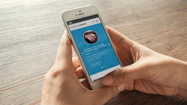 M&atilde;os segurando um celular sobre uma mesa. Na tela do celular est&aacute; aberto um site do modelo hotsite do Matriz.