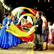 Grupo de casais vestidos com roupas tradicionalistas do Rio Grande do Sul segurando e sacudindo tecidos nas cores amarela, verde e vermelha.