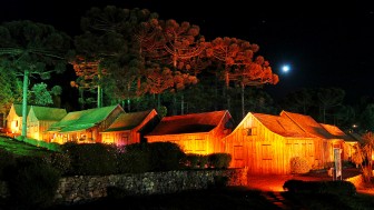 Casas t&iacute;picas de madeira da serra ga&uacute;cha iluminadas &agrave; noite, sob as arauc&aacute;rias, em noite de lua cheia.